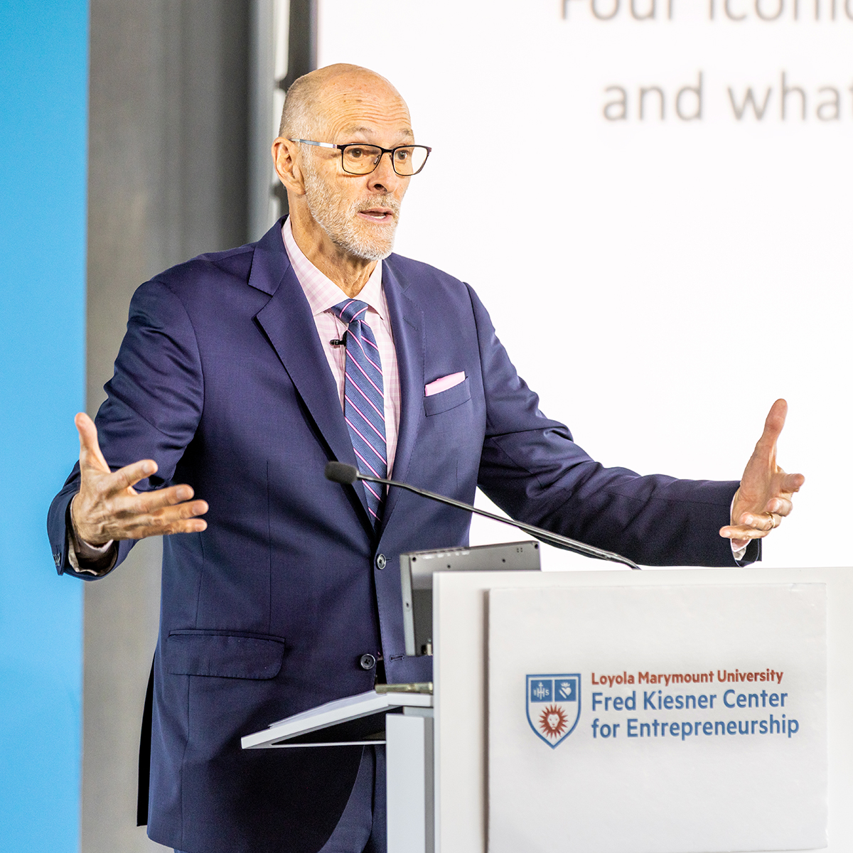 A guest speaker standing behind a podium mid-speech with their arms extended.